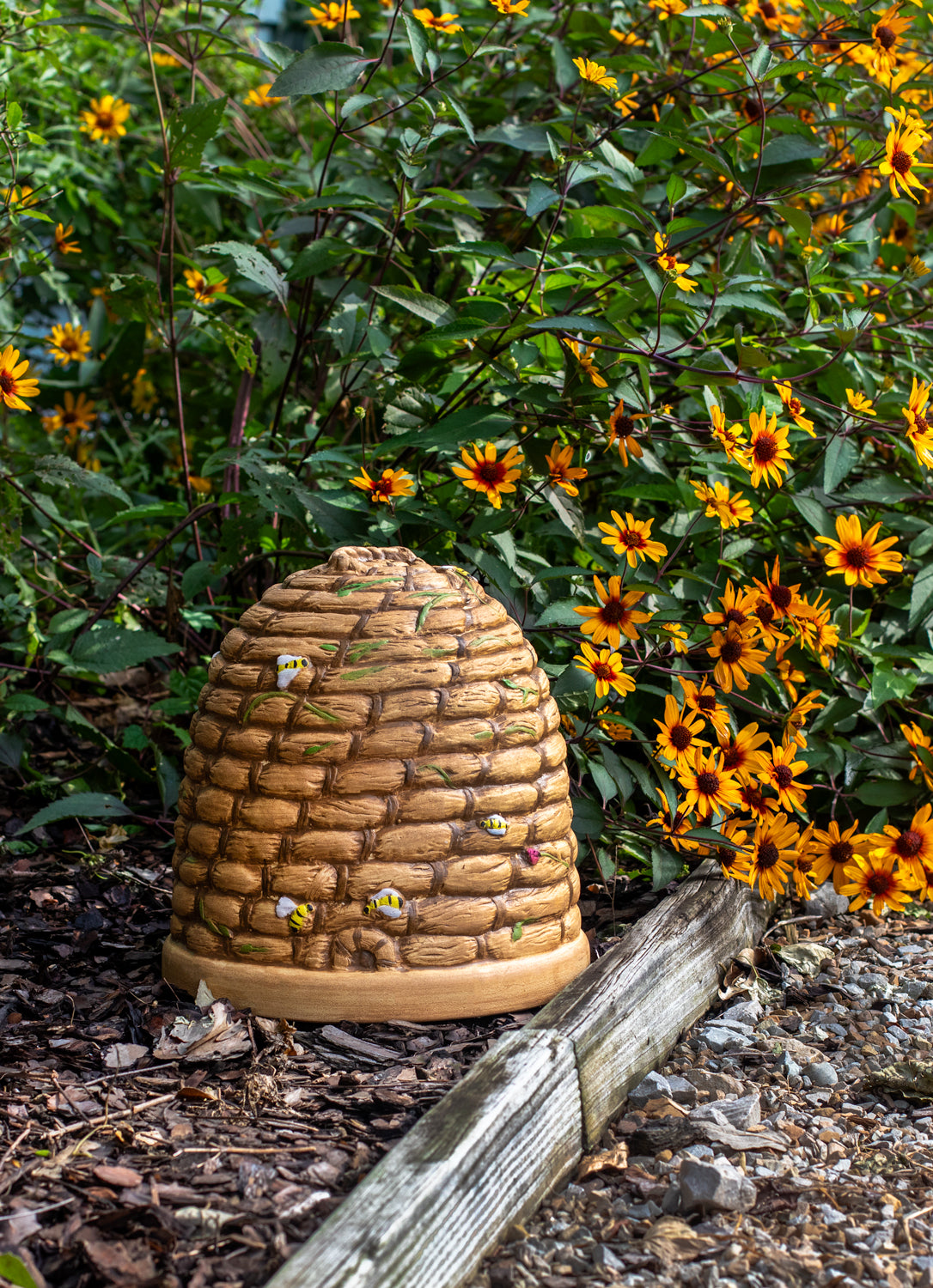 Beehive-shaped garden accessory surrounded by greenery and yellow flowers in a bed of mulch next to a stone pathway