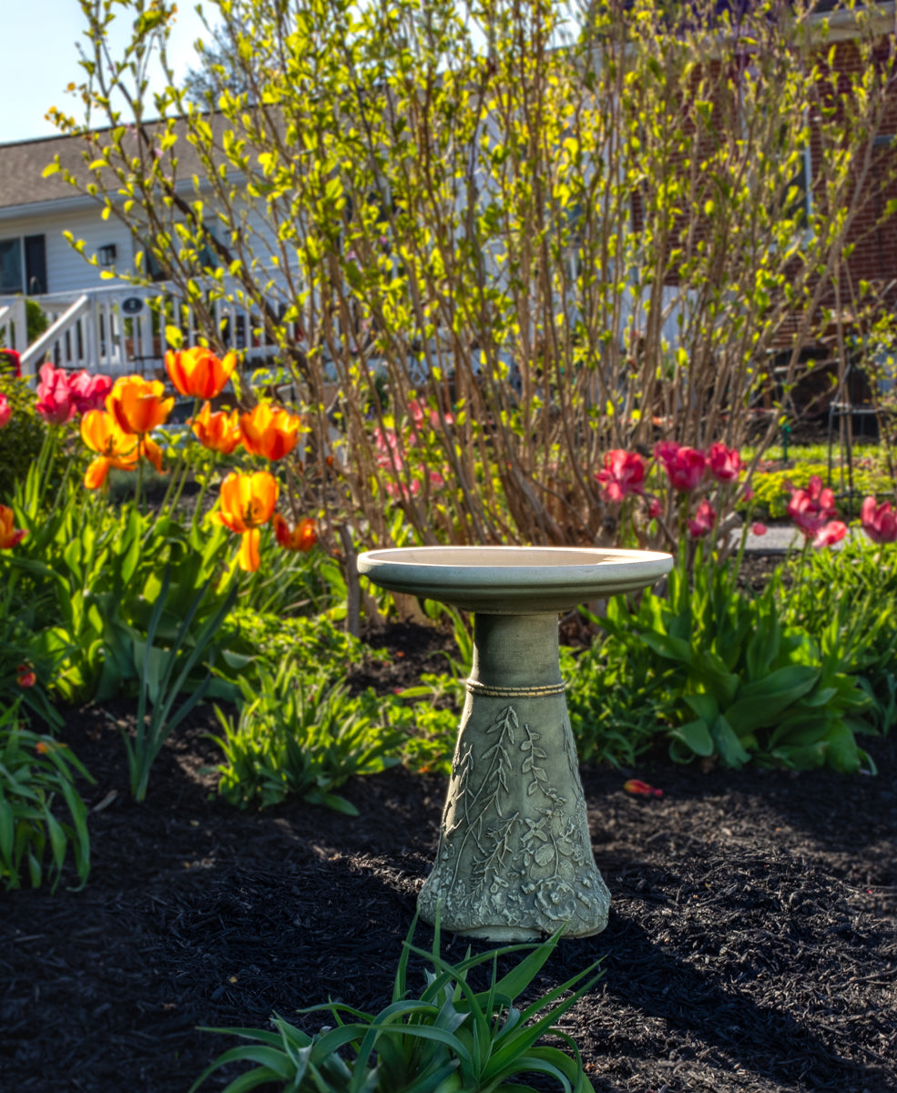 shorter ceramic birdbath set set in a flower bed filled with brightly colored flowers and large bush