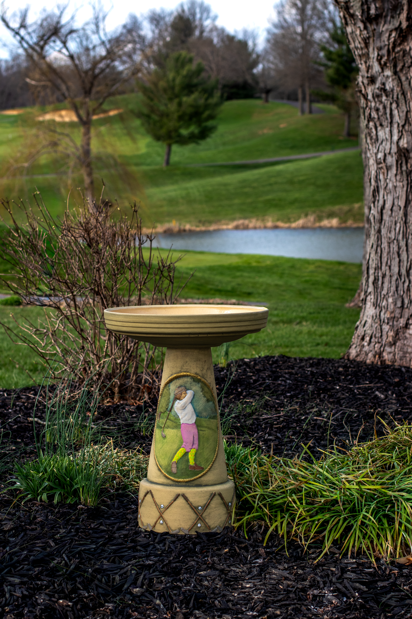 Decorative bird bath with a golf-themed design in a garden setting with grass and trees.
