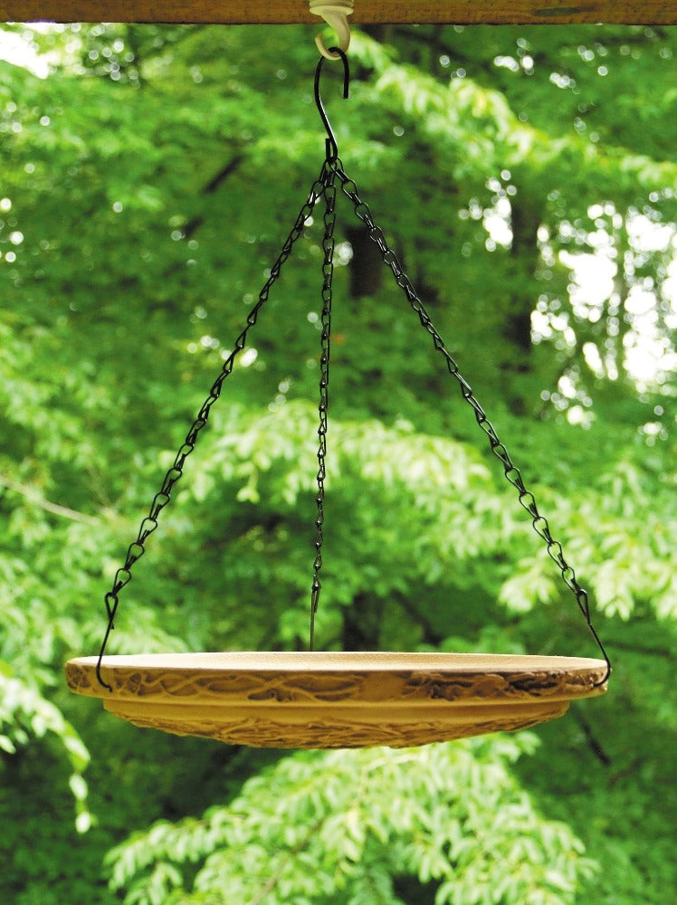 A beige, hanging nest birdbath feeder made of clay, suspended by chains against a backdrop of green foliage.