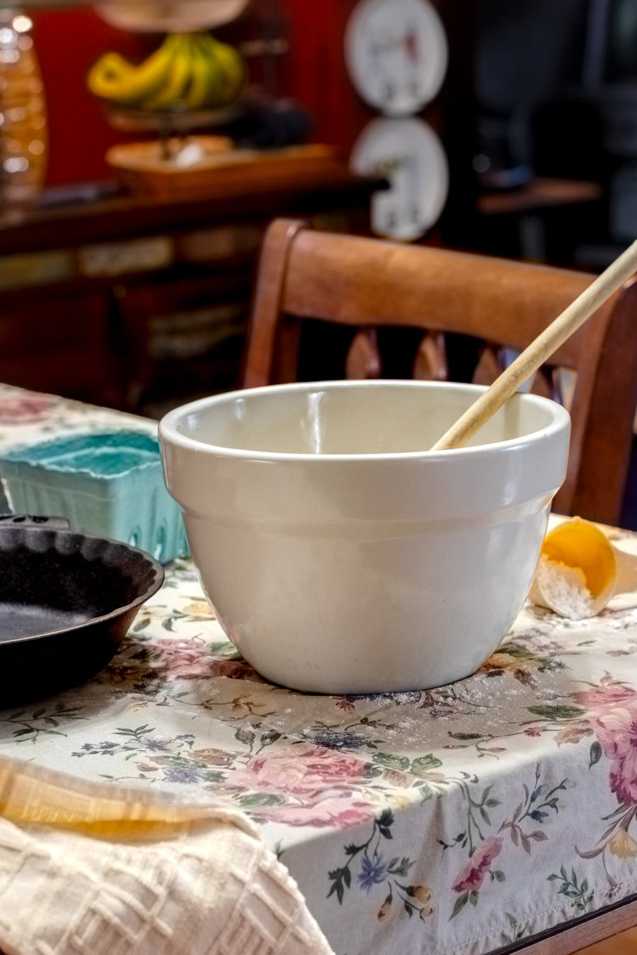 White Ceramic mixing bowl with a wooden spoon on a floral tablecloth next to a cast iron pie dish and dish towel