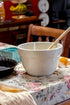 White Ceramic mixing bowl with a wooden spoon on a floral tablecloth next to a cast iron pie dish and dish towel
