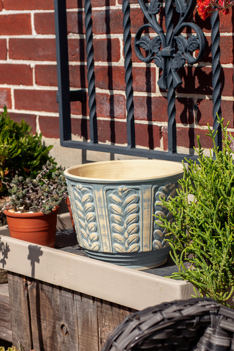 A large light blue and beige ceramic planter with embossed leaf patterns sits on a wooden planter box in an outdoor garden setting, with a brick wall, wrought iron gate, and potted plants in the background.